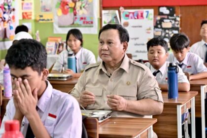 Man in a khaki uniform seated at a desk in a classroom, surrounded by students in white shirts and ties.