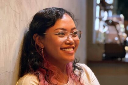 Smiling woman with glasses sits against a textured wall in a warm indoor setting.