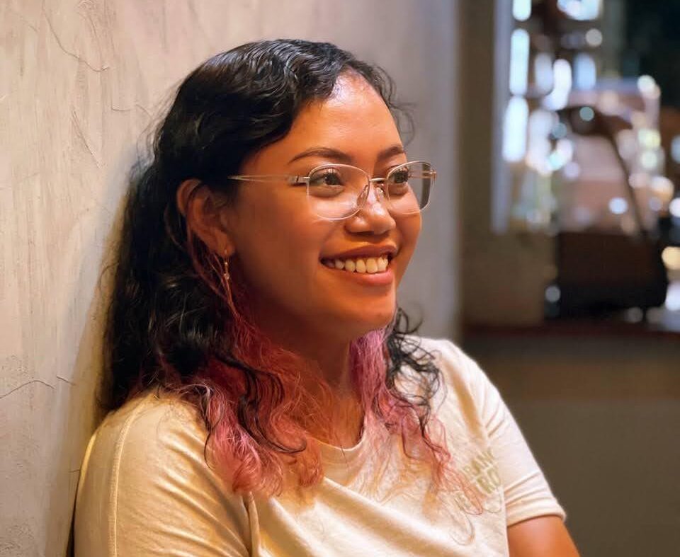 Smiling woman with glasses sits against a textured wall in a warm indoor setting.